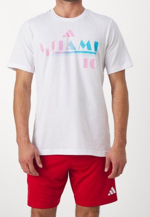Mand iført hvid T-shirt med teksten "Miami 10" i pink og blå samt røde shorts med Adidas-logo på venstre ben.