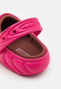 Crocs rosa in materiale gommoso, con una superficie testurizzata e un interno in peluche marrone. Presenta una fascia con il logo e fori di ventilazione.