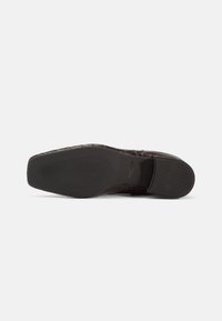 Semelle en cuir marron avec un motif texturé, présentant une base en caoutchouc noir lisse et un piquage détaillé le long des bords.