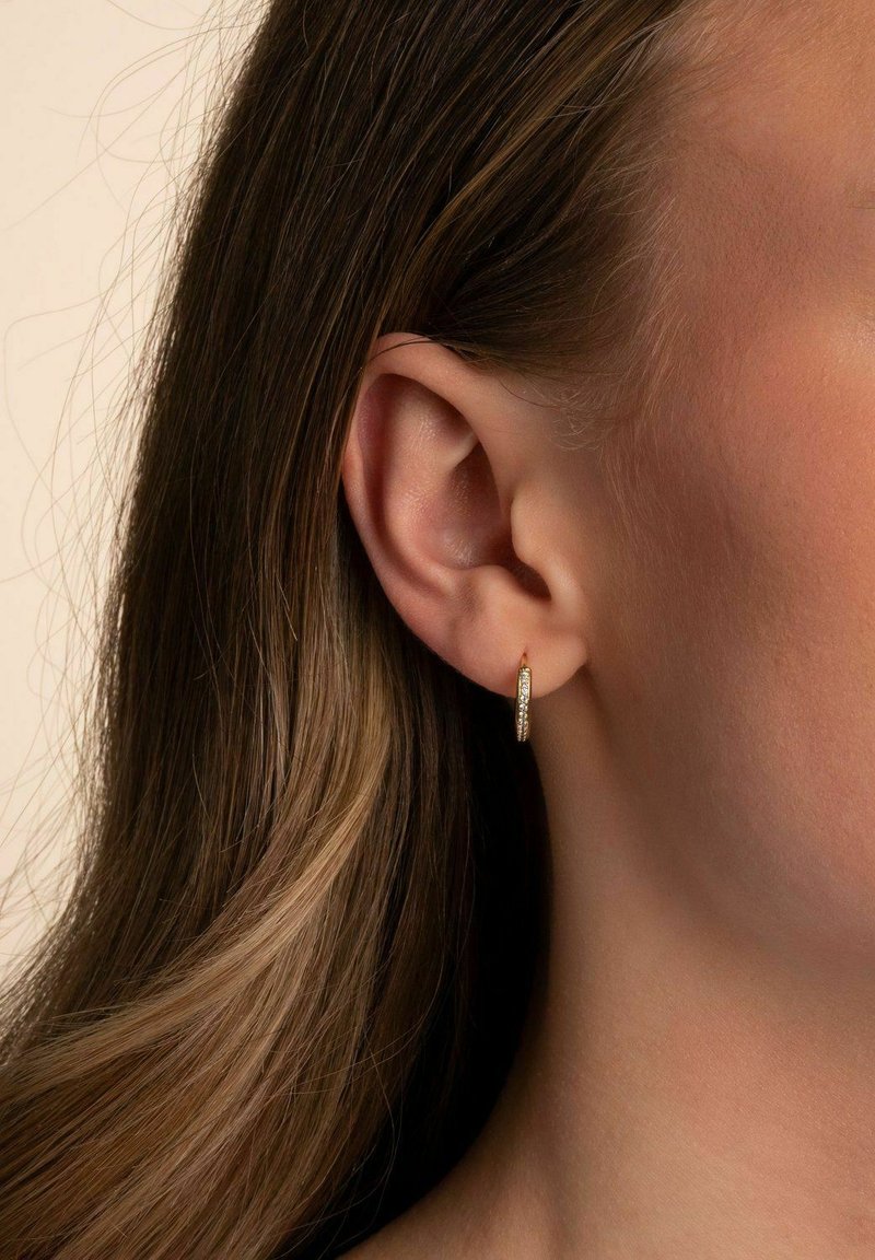 Guldörhänge i form av en ring med en strukturerad yta och små inslagna klara stenar. Designen är cirkulär och reflekterar ljus på olika sätt.