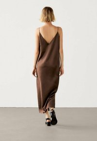 Brązowa, bezrękawnikowa sukienka z dekoltem w kształcie litery V, z lejącym się materiałem, o długości do kostek, noszona z czarnymi sandałami na słupku.