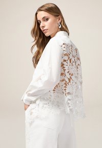 Weißes Hemd mit einem Rückteil aus floraler Spitze, langen Ärmeln und Knopfmanschetten. Verfügt über einen Kragen und ein lässiges Design. Glatte Stoffoberfläche.