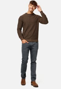 Brązowy sweter z teksturowanej dzianiny z okrągłym dekoltem, zestawiony z szarymi dżinsami i brązowymi botkami za kostkę, na modelce stojącej w swobodnej pozie.