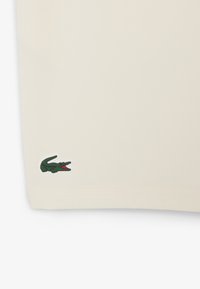 Camisa beige de manga corta, con un logotipo de cocodrilo verde en el dobladillo inferior izquierdo y una suave textura de algodón.