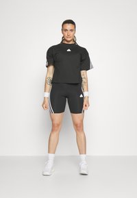 Μαύρο κοντό μπλουζάκι και ταιριαστά σορτς με ψηλή μέση, και οι δύο με λευκές τριές ρίγες, συμπληρωμένα από λευκά βραχιόλια και αθλητικά παπούτσια.