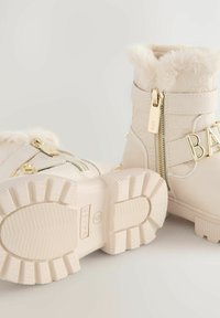 Stivali invernali beige con colli soffici e dettagli dorati. Presentano cerniere laterali doppie e suole testurizzate. Design elegante e alla moda.