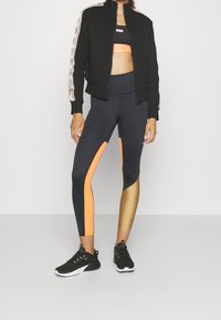 Chaqueta negra con cremallera y franja lateral beige, top ajustado negro, leggins negros y naranjas, y zapatillas deportivas negras. Texturas suaves, colores vibrantes.