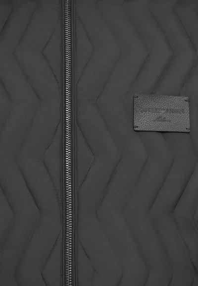 Black quilted jacket fabric with zigzag stitch pattern, central silver zipper, and a black rectangular Emporio Armani Milano label.
