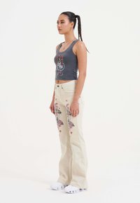 Débardeur gris avec impression graphique, jean beige évasé avec des motifs floraux et un motif cœur, associé à des chaussures de sport blanches.