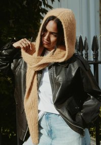 Beige Wollschal mit integrierter Kapuze, kombiniert mit einer schwarzen Lederjacke und einem weißen Hemd. Die Textur wirkt weich und warm. Lässiges Outfit.