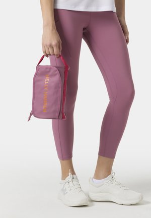 Persona que lleva leggings malva y zapatillas blancas, sosteniendo una bolsa malva de Helly Hansen con asas rojas a su lado.
