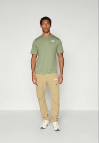 Hombre de pie con una camisa de manga corta verde oliva, pantalones cargo beige y zapatillas blancas frente a un fondo claro y liso.