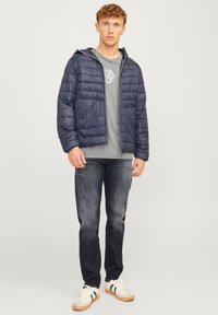 Merikadun puffer-takki vaakasuoralla tikatulla kuvioinnilla, harmaa logo-t-paita, tummat farkut ja valkoiset lenkkarit vihreillä korostuksilla.