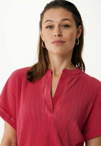 Červený žebrovaný top s krátkým rukávem, který má výstřih do V a malou légou, zdůrazňující jemnou strukturu a uvolněný střih.