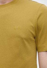 Gros plan d'un T-shirt jaune moutarde à manches courtes avec un petit texte brodé « BOSS » sur la poitrine.