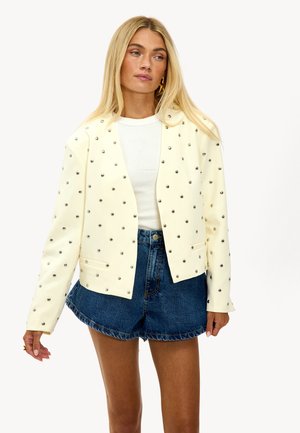 Blonde vrouw in een crèmekleurige jas met zilveren studs, witte top en high-waisted spijkerbroekshorts, poserend tegen een effen achtergrond.