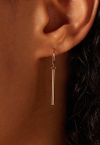 Boucle d'oreille en métal doré en forme de créole avec une longue barre verticale pendante, surface lisse et design minimaliste, mise en valeur sur une oreille.