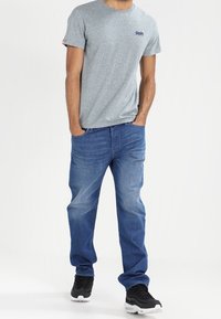 Harmaa lyhythihainen t-paita logolla, siniset farkut ja mustat tennarit valkoisilla yksityiskohdilla, seisoo vaaleaa taustaa vasten.