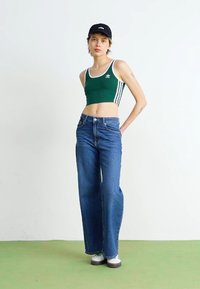 Grön ärmlös croptop med vita ränder, matchad med högmidjade blå vida jeans. Modellen bär en svart keps och vita sneakers.
