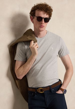 Άνδρας με σγουρά κόκκινα μαλλιά και γυαλιά ηλίου φοράει γκρι μπλουζάκι, σκούρα τζιν με καφέ ζώνη και ρολόι, κρατώντας ένα καφέ σακάκι στον ώμο.