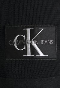 Černá látková nášivka s nápisem "CALVIN KLEIN JEANS" ve stříbrné výšivce, s výrazným logem "CK". Texturované pozadí dodává detail.