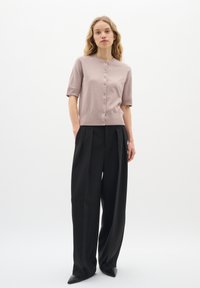 Helles rosa, kurzärmliges Cardigan mit rundem Halsausschnitt und Knopfverschluss, kombiniert mit maßgeschneiderten, hochgeschnittenen schwarzen Hosen und spitzen schwarzen Schuhen.