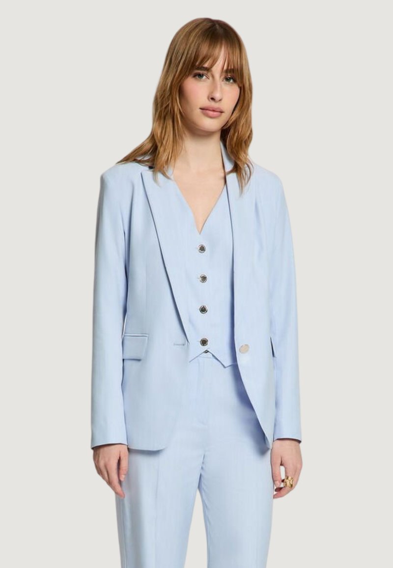 Femme aux cheveux châtain clair portant un blazer bleu clair sur mesure, un gilet assorti boutonné et un pantalon, debout devant un fond uni.