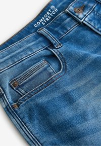 Primer plano de un bolsillo de jeans de mezclilla azul con costuras detalladas, remaches metálicos, botón y etiqueta de "Comfort Stretch" cerca de la cinturilla.
