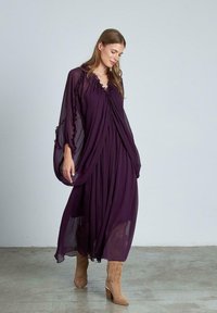 Długa fioletowa sukienka z szyfonu z falbaniastym dekoltem i rękawami, o zwiewnym kroju, zestawiona z brązowymi botkami. Gładka tekstura i lekki materiał.
