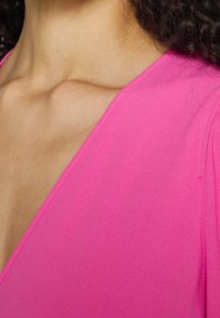 Fuchsia V-Ausschnitt-Top mit glatter Textur, das eine saubere Kante und leichte Nähte entlang des Halsauschnitts und der Ärmel aufweist.