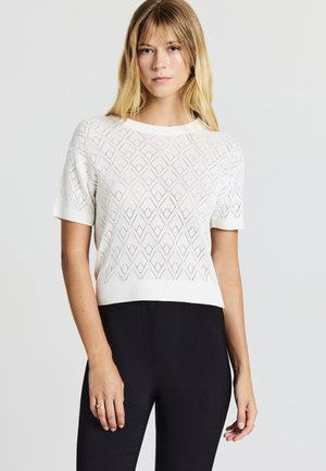 Frau mit blonden Haaren, die einen weißen kurzärmligen Strickpullover mit Rautenmuster und schwarze Hosen vor einem einfarbigen Hintergrund trägt.