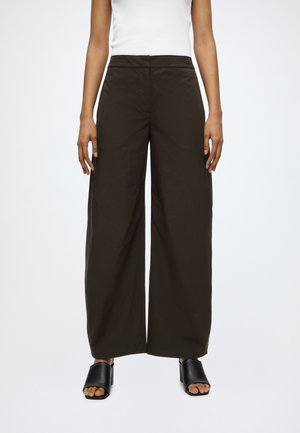 Femme portant un pantalon large marron foncé, un top blanc sans manches et des sandales à talons noires ouvertes, debout, les bras détendus.