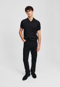 Camiseta tipo polo negra de manga corta con tapeta de tres botones, combinada con jeans negros y zapatos de ante negros, sobre un fondo neutro.