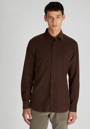 Man in een donkerbruin corduroy overhemd met knopen en beige broek, staand tegen een effen lichtgrijze achtergrond.