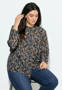Frau mit langen, dunklen Haaren, die eine blau-braun-schwarz gemusterte Bluse und blaue Jeans trägt, sitzt auf einem Hocker vor einem weißen Hintergrund.
