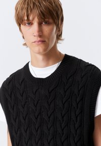 Fekete kötött mellény texturált kábelmintával, széles karöltőkkel és kerek nyakkivágással, fehér póló fölött viselve.