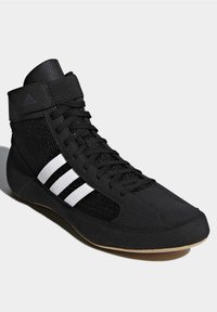 Baskets montantes noires Adidas avec des bandes blanches, des empiècements en mesh, des lacets et une bride à la cheville sur fond gris clair.