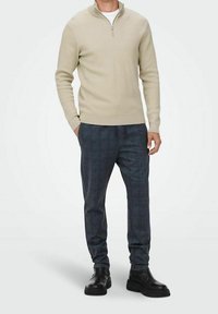 Beige striktrøje med lynlås ved halsen i en ribbet tekstur kombineret med ternede mørkeblå bukser. Modellen bærer sorte ankelstøvler. Enkel, moderne pasform.