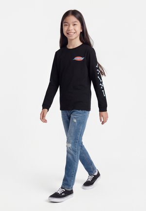 Fată zâmbitoare care merge, purtând un tricou negru cu mâneci lungi cu logo și modele cu oase pe mâneci, blugi albaștri rupți și adidași negri.