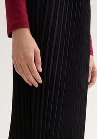 Schwarzer plissierter Rock mit weicher Textur, der neben einer Hand gezeigt wird, die auf dem Stoff ruht. Lange schwarze Falten, die vertikal verlaufen, betonen das Design.