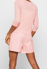 Personne portant un haut en maille rose clair à motif chevron et un short rose assorti, vue de dos avec un bras légèrement plié.