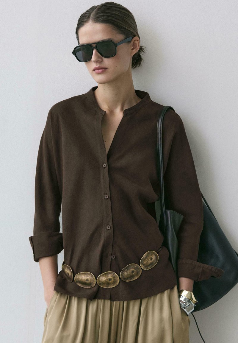 Femme portant des lunettes de soleil foncées, une chemise marron boutonnée, une jupe beige, une large ceinture ovale, un bracelet en or et un sac à main noir.