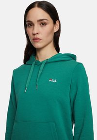 Sudadera de forro polar verde con bolsillo canguro, capucha ajustable por cordón y logo bordado de FILA en el pecho, con ojales de tono plateado.