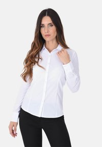 Camicia bianca a maniche lunghe con colletto, abbottonatura frontale e vestibilità slim. Tessuto di cotone liscio, polsini piatti e orlo curvato in vita.