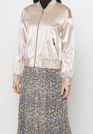 Femme portant une veste bomber beige clair brillante avec des poches zippées sur un haut noir et une jupe à motif floral avec des fleurs violettes et jaunes.