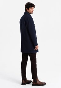 Cappotto di lana blu navy con colletto, chiusura a un bottone e tasche laterali. Abbinato a pantaloni marroni e stivali alla caviglia marroni scuri.