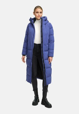 Frau trägt einen langen blauen Steppmantel mit Kapuze, weißen Rollkragenpullover, schwarze Jeans und schwarze Stiefeletten, die auf einem schlichten hellen Hintergrund steht.