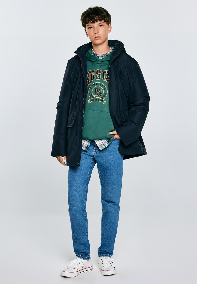 Mornarska puhovka s kapuco, zelena majica s logotipom, karirana srajca, svetlo modri skinny jeans in bele superge. Ohlapna večslojna kombinacija.
