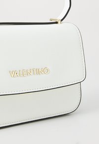 Borsa a tracolla in pelle bianca con rifiniture nere, logo dorato "VALENTINO" e design a patta strutturato. Accentuata da hardware dorato e cuciture.