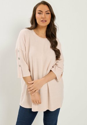 Kvinde iført en løs, lys beige top med trekvartærmer og knappedetaljer, kombineret med mørkeblå jeans, stående mod en ensfarvet baggrund.